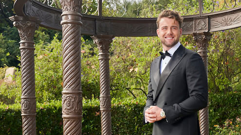 Man in a black tuxedo smiling outdoors, standing near ornate pillars and greenery, with an elegant garden gazebo structure in the background.