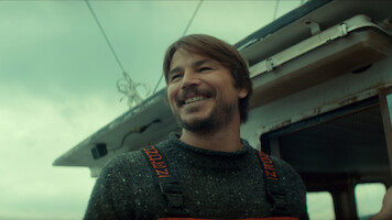 Smiling man with brown hair wearing a sweater and overalls stands on a boat under a cloudy sky, with fishing equipment and parts of the boat visible in the background.