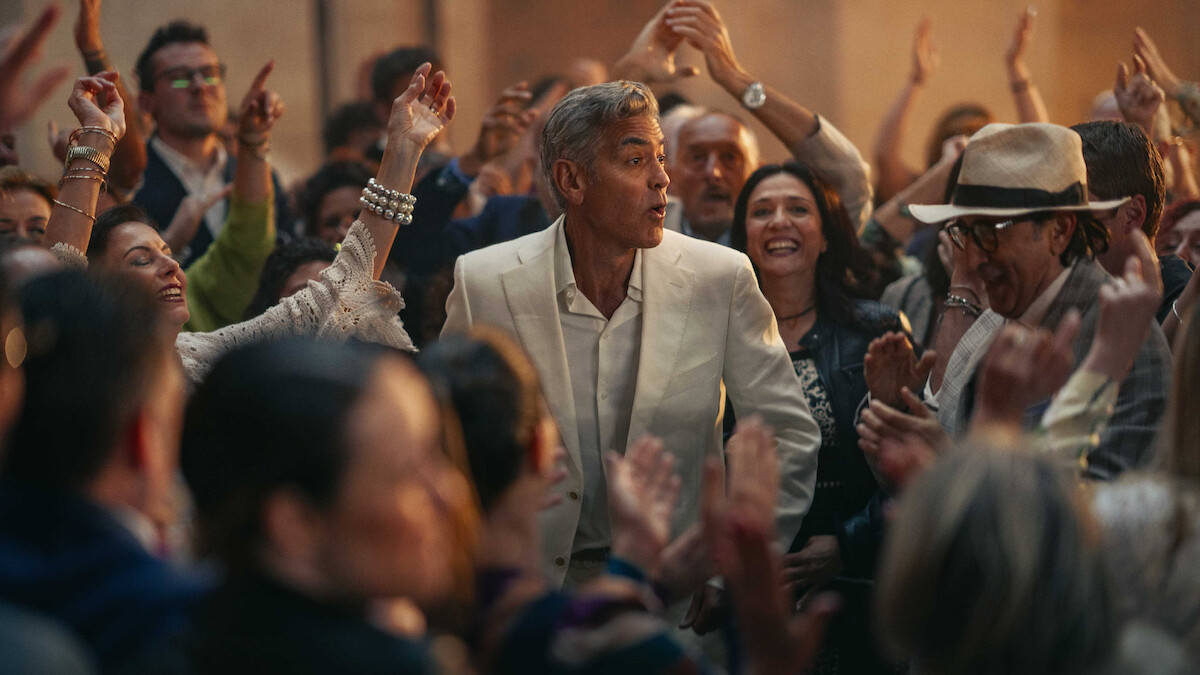 A man in a white suit stands surrounded by a lively crowd at a festive gathering, with smiling faces and raised hands creating a joyful, celebratory atmosphere.