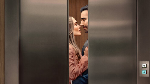 A man and woman standing closely, facing each other romantically inside an elevator as the doors begin to close, suggesting a private or intimate moment in a modern building setting.