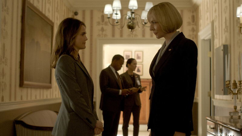 Two women in business suits face each other in a well-lit hallway with elegant decor; two other people are talking in the background, suggesting a formal or professional setting.