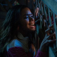 A woman in distress gripping a wooden lattice fence, emotional and shouting in a dimly lit environment, suggesting a tense or dramatic situation.