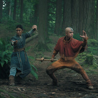 Two young people in fantasy warrior costumes pose in martial arts stances with focused expressions in a lush, green forest surrounded by tall trees and ferns, suggesting an adventurous or mystical setting.