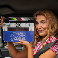Smiling woman in a car holding a film clapperboard labeled "this summer will be different" wearing a red-checkered shirt and seatbelt, filming scene in bright daylight.