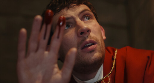 A worried man in a red robe holds up his hand with blood on his fingers, looking upwards in a dimly lit, stone-walled setting, creating a tense and dramatic mood.