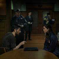 A man and a woman sit across from each other at a table in a dimly lit room, with police officers and investigators working in the background, suggesting an interrogation or police investigation scene.