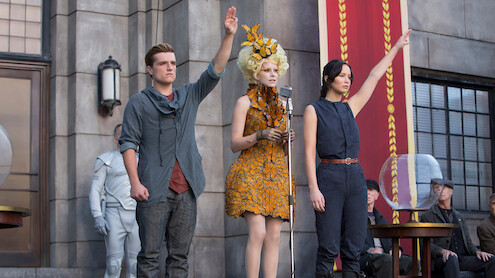 Three people stand in front of a stone building, two raising one hand; one wears a grey shirt, another a sleeveless outfit, and the third is in an elaborate orange dress at a microphone. Others are seated nearby, and a large banner hangs.