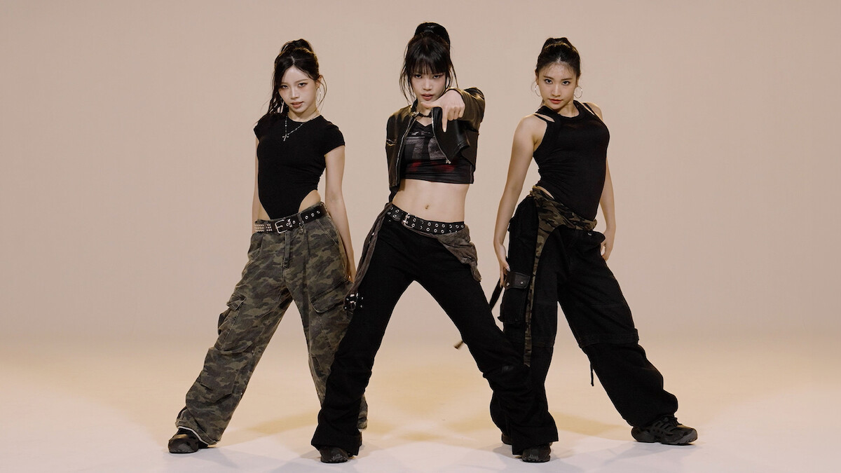 Three women in black and camo outfits pose confidently in a studio with a plain beige background, suggesting a dance or performance setting.