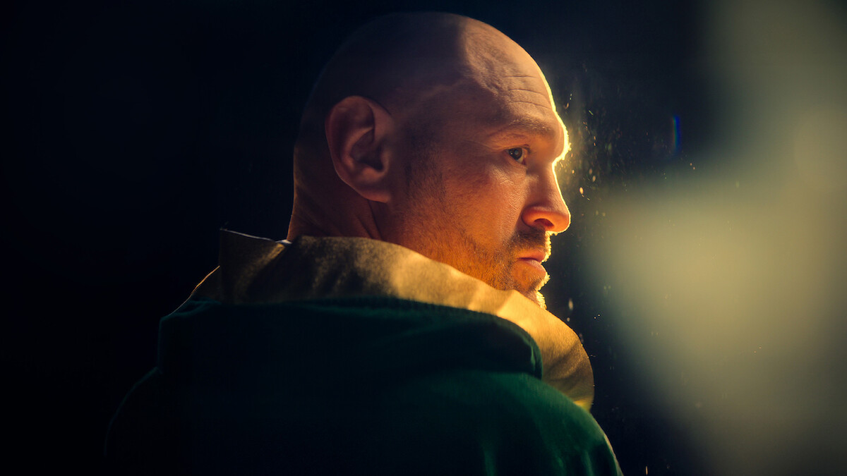 A bald man in a hooded jacket stands in a dimly lit environment, looking over his shoulder towards a warm, soft light source that highlights one side of his face against a dark background.