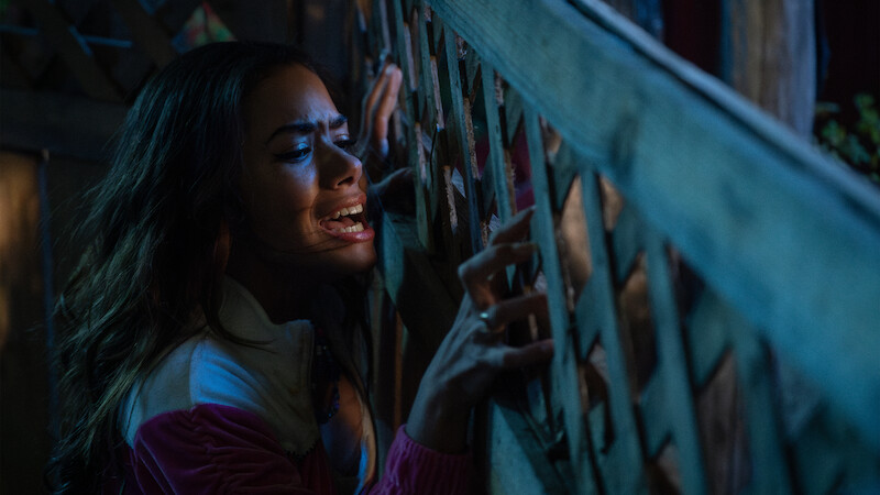 A distressed woman grips a wooden lattice fence at night, illuminated by dramatic lighting, expressing fear or desperation in an outdoor setting.