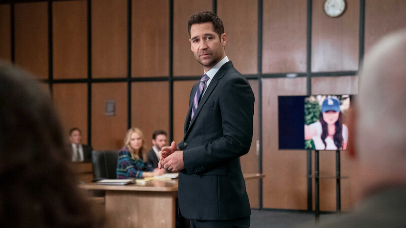Man in suit stands in courtroom, addressing others. People sit at a table in the background. A digital screen shows a woman’s photo. Wood panel walls and a clock indicate a formal court setting.