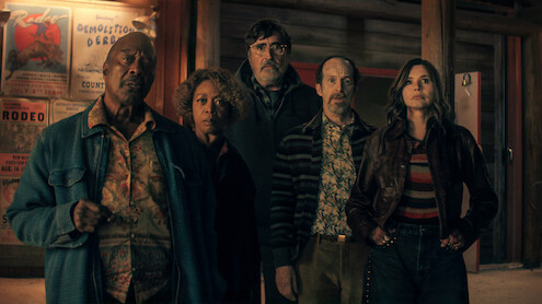Five people stand indoors in front of wooden walls with posters, looking intently ahead, wearing retro-style clothing in a dimly lit setting suggestive of a bar or lodge.