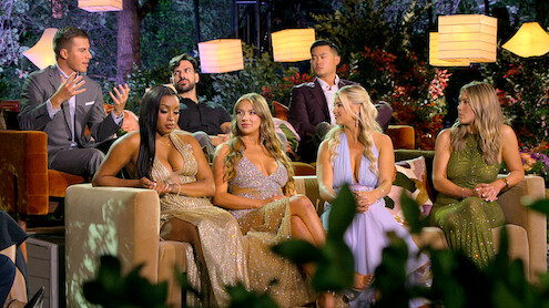 Six people in formal attire sit on couches outdoors at night, surrounded by lush greenery and hanging lamps, engaged in conversation with a vibrant, glamorous atmosphere.