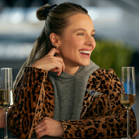 Smiling woman in a leopard print jacket and gray hoodie sitting at a table with two glasses of champagne, in a modern indoor setting with soft lighting and blurred green plants in the background.