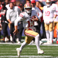 San Francisco 49ers football player in uniform running with the ball on a sunny field during a game, with teammates and spectators in the background.