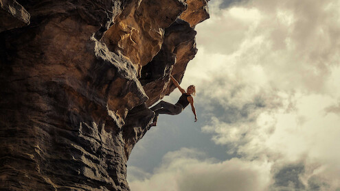 A rock climber scaling a steep, overhanging cliff outdoors under a cloudy sky, demonstrating strength and balance in a dramatic natural setting.