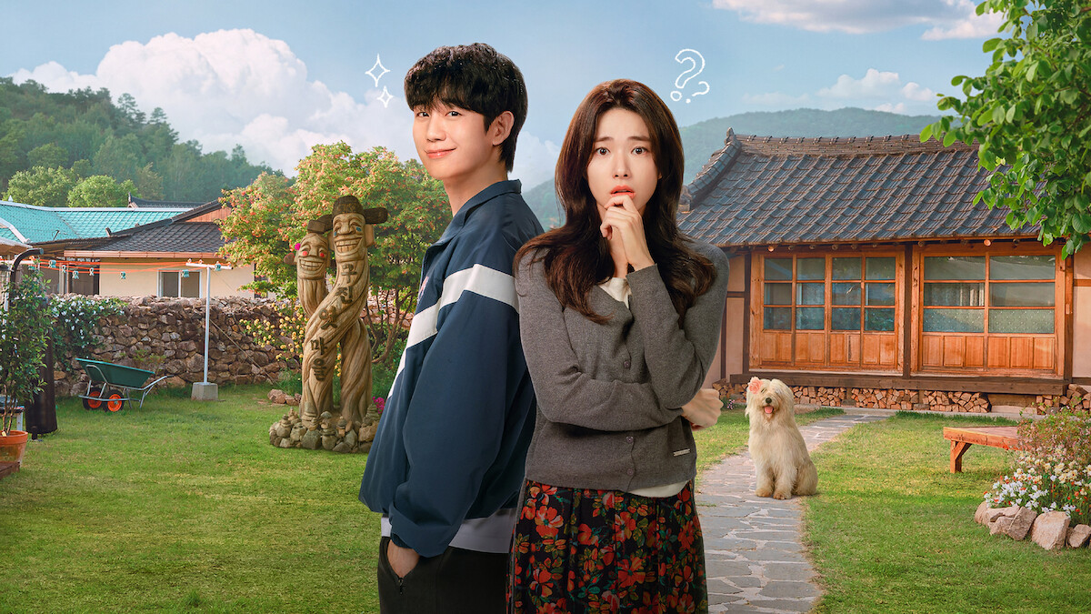 A young man and woman stand in a sunny, rural yard with traditional wooden house, lawn, garden statues, and a fluffy dog, mountains in the background, blue sky with clouds, both subjects displaying thoughtful and playful expressions.