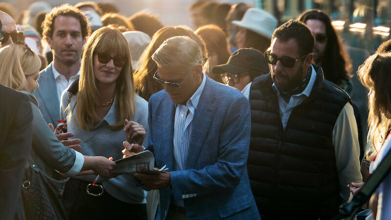 A man in a blue suit signs autographs in a crowded outdoor setting at sunset. People around him wear sunglasses and casual clothing, creating an excited and lively atmosphere.
