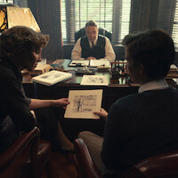 Three people in an office: a man behind a desk and two others sitting across, showing sketches. The setting has classic decor, sunlight filtering through blinds, and papers and drawings spread out, suggesting a serious discussion or review.