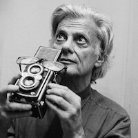 Older man with gray hair in a collared shirt holding a vintage twin-lens reflex camera, gazing thoughtfully upward in an indoor setting with neutral background. Black and white photo, studio light ambiance.