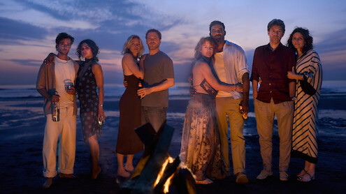 Four couples stand together on a beach at sunset near a campfire, dressed in casual evening clothes, some holding drinks, ocean and colorful sky in the background.