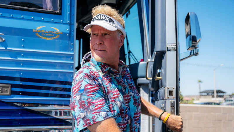 Man in a tropical shirt and visor stands at the doorway of a blue luxury bus on a sunny day, holding the door handle, with a fence and clear sky in the background.
