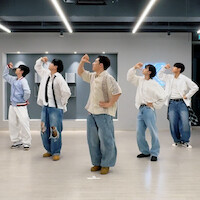  Five people in casual clothing pose in a dance studio with light wooden floors, modern decor, and a neon sign on the wall, all striking the same choreographed dance pose facing left.