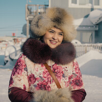 Woman smiling outdoors in snowy environment, wearing a floral coat and fur hat, with houses and snow-covered ground in the background, suggesting a cold winter setting.