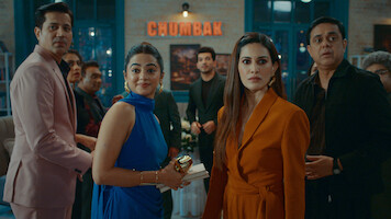 A group of well-dressed people at an indoor gathering or party, looking toward the camera with expressions of surprise or curiosity; modern decor and a sign reading "CHUMBAK" in the background.