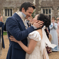 A couple dressed in Regency-era wedding attire embrace outdoors, surrounded by clapping guests in formal period clothing, with a stone building in the background.