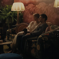 A couple sits closely together on a floral sofa with a small dog in a cozy, dimly lit living room with warm lighting, plants, patterned walls, and a table beside them.