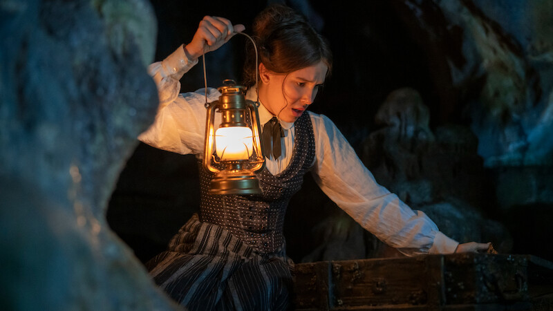 A woman in period clothing holds a lit lantern while exploring a dark, rocky cave, leaning over an old wooden chest, suggesting a search or discovery in a mysterious setting.