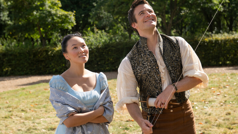 A woman and man in period costumes stand outside on a sunny day, smiling and looking up while the man flies a kite.