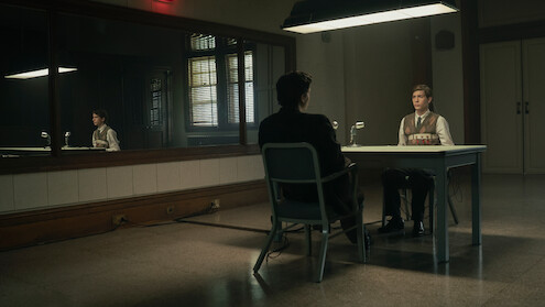 Two people sit across from each other at a table in a dimly lit interrogation room, with a large mirror on the wall reflecting the scene and a microphone on the table, creating a serious and tense atmosphere.