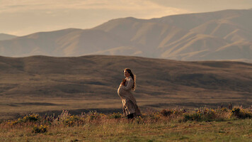 Pregnant woman in a long dress stands in a grassy field with wildflowers, set against a backdrop of rolling hills and mountains during golden hour, creating a serene and natural outdoor scene.