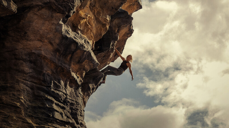 Person rock climbing on a steep, overhanging cliff with dramatic rock formations, reaching outward against a cloudy sky, outdoors in a rugged natural environment.