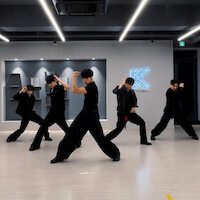 Five dancers in black outfits pose mid-routine in a modern dance studio with bright ceiling lights and a glowing K logo on the wall, light wooden floors, and geometric wall decor in the background.