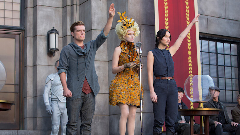 Three people stand in front of a stone building, two raising one hand; one wears a grey shirt, another a sleeveless outfit, and the third is in an elaborate orange dress at a microphone. Others are seated nearby, and a large banner hangs.