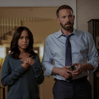 Two people, a concerned woman in a sweater and a serious man in a dress shirt and tie, stand in a modern home interior with staircase in the background, appearing tense or worried.