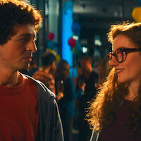 A man and woman talking at a lively indoor party with colorful balloons and people socializing in the background, warm lighting.