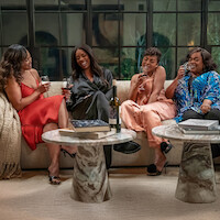 Four women sit on a modern sofa in a stylish living room, laughing and enjoying glasses of wine. Two round marble tables with books and a bottle of wine sit in front of them. Large windows and cozy decor complete the setting.