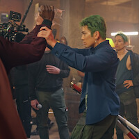 Actor with green hair in a blue shirt poses with a prop sword on a film set, rehearsing a scene with a crew member holding a water bottle; other crew and film equipment visible in the background of an industrial setting.