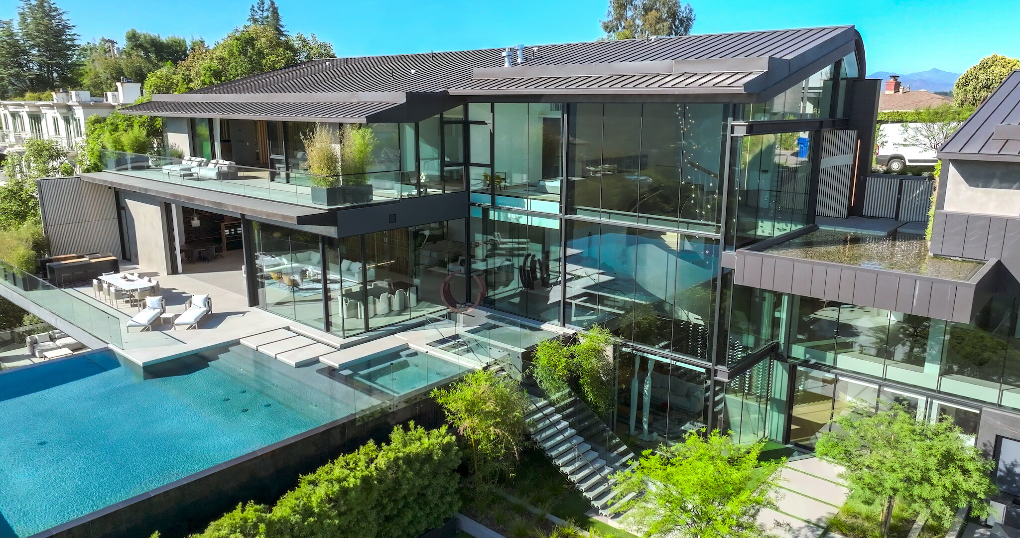 Buying Beverly Hills key art- A photo of a mansion with large glass windows, a pool, in ground jacuzzi, and stairs leading down to a garden area.