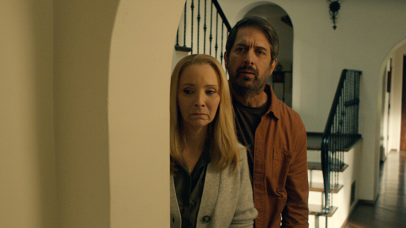 Ray Romano as Paul and Lisa Kudrow as Lydia hide behind a doorway with worried expressions in Season 1 of 'No Good Deed'