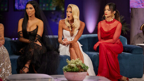 Three women in elegant dresses sit on a blue couch in a stylish, dimly lit room. The mood is tense and dramatic, with one woman appearing shocked and the others looking serious.