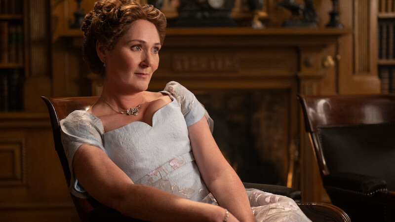 A woman in a light blue period dress sits in a wooden chair in an ornate library or study, with wood paneling, bookshelves, and a decorative fireplace in the background, looking thoughtful.