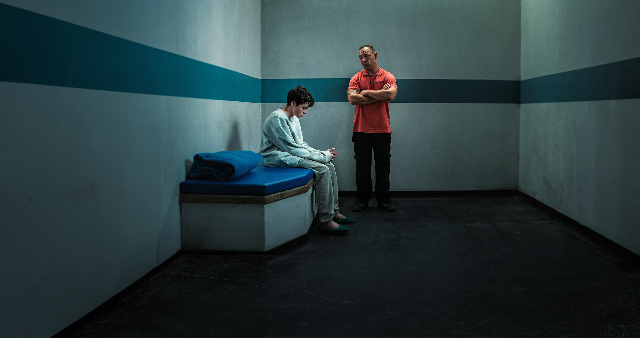 Adolescence key art - A man in an orange shirt is looking down on a teenager who looks despondent while sitting on the edge of his bed. 