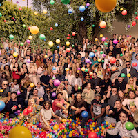 Large group of people gathered outdoors under trees, posing together while colorful balloons float around them, creating a festive, cheerful atmosphere in daylight.