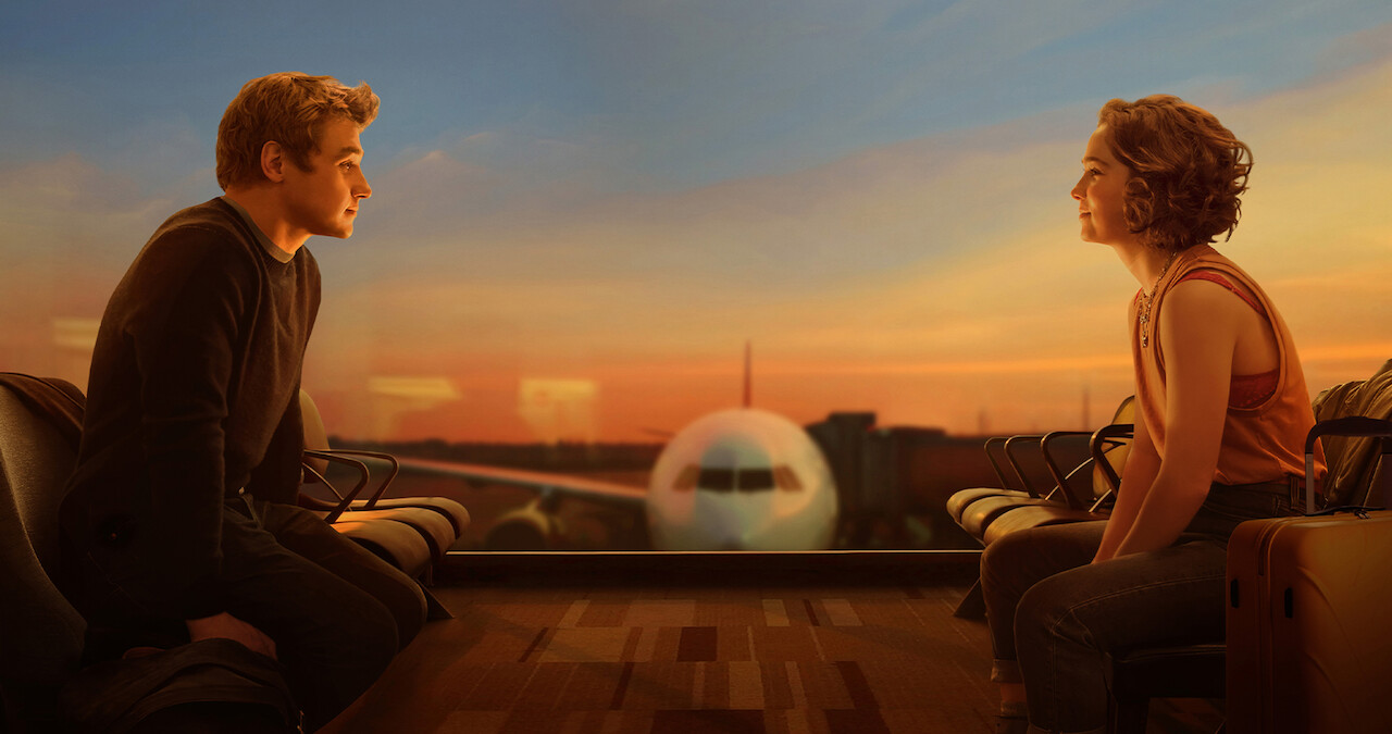 Ben Hardy and Haley Lu Richardson sit opposite one another on an airport terminal floor, with a plane between them outside the window.