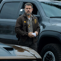 A police officer with a badge and notepad stands between parked vehicles outdoors, appearing focused and alert, with overcast lighting and paperwork in hand.